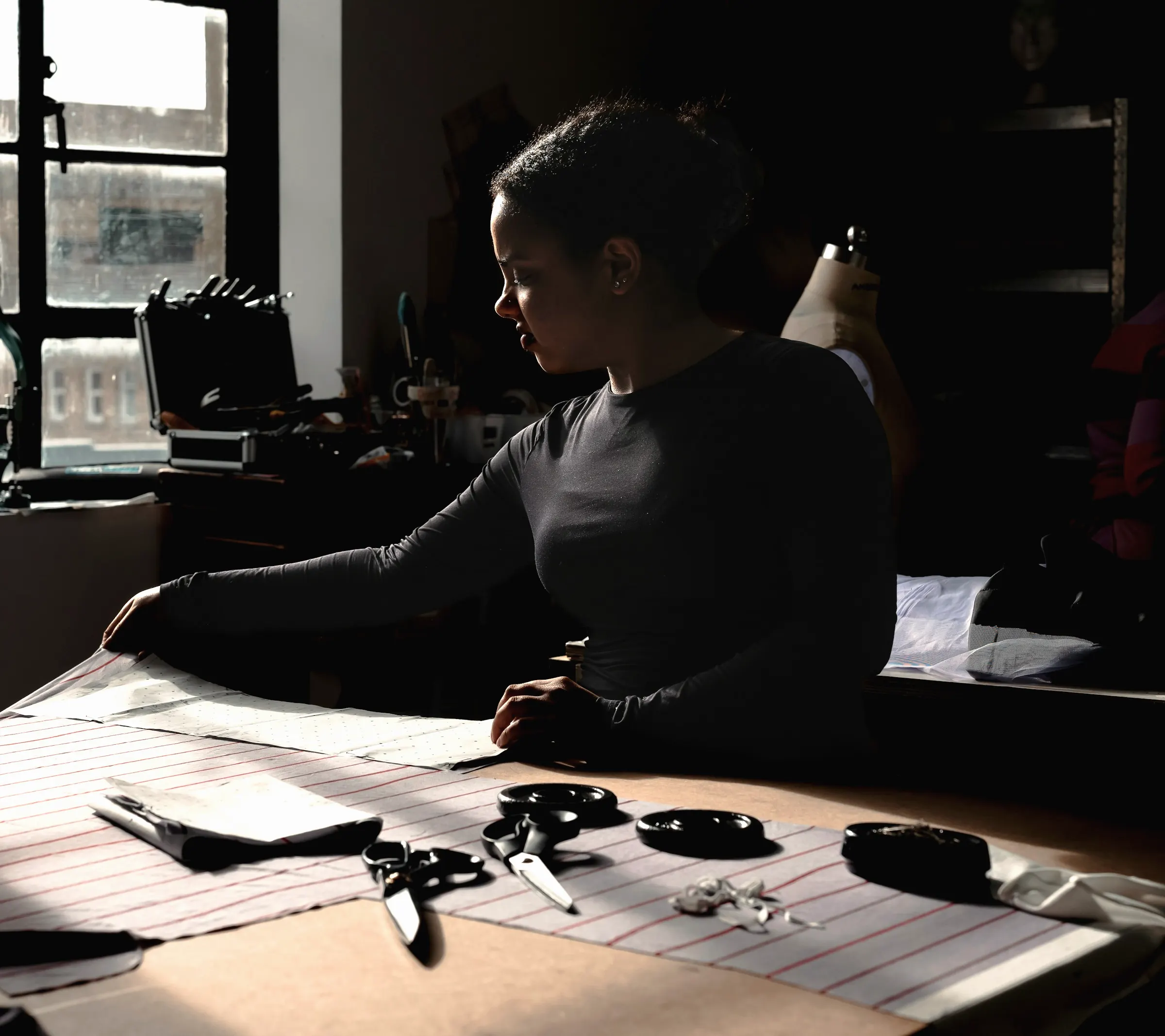 A maker working at a large pattern-cutting table in a moody, window-lit studio, scissors and tools laid out on striped fabric
