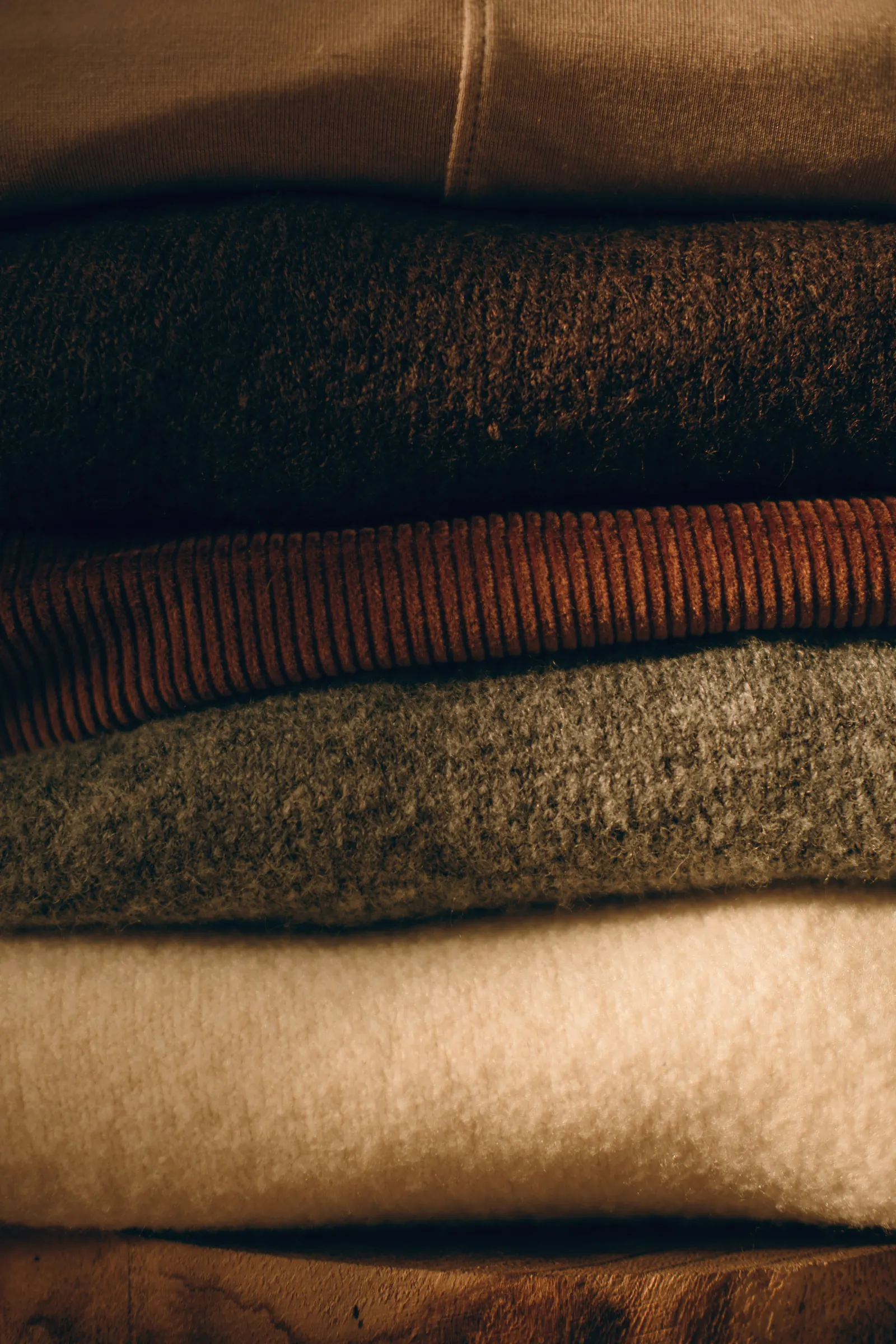 A tall stack of folded sweats and knits in espresso, rust, olive and cream, lit by low warm light