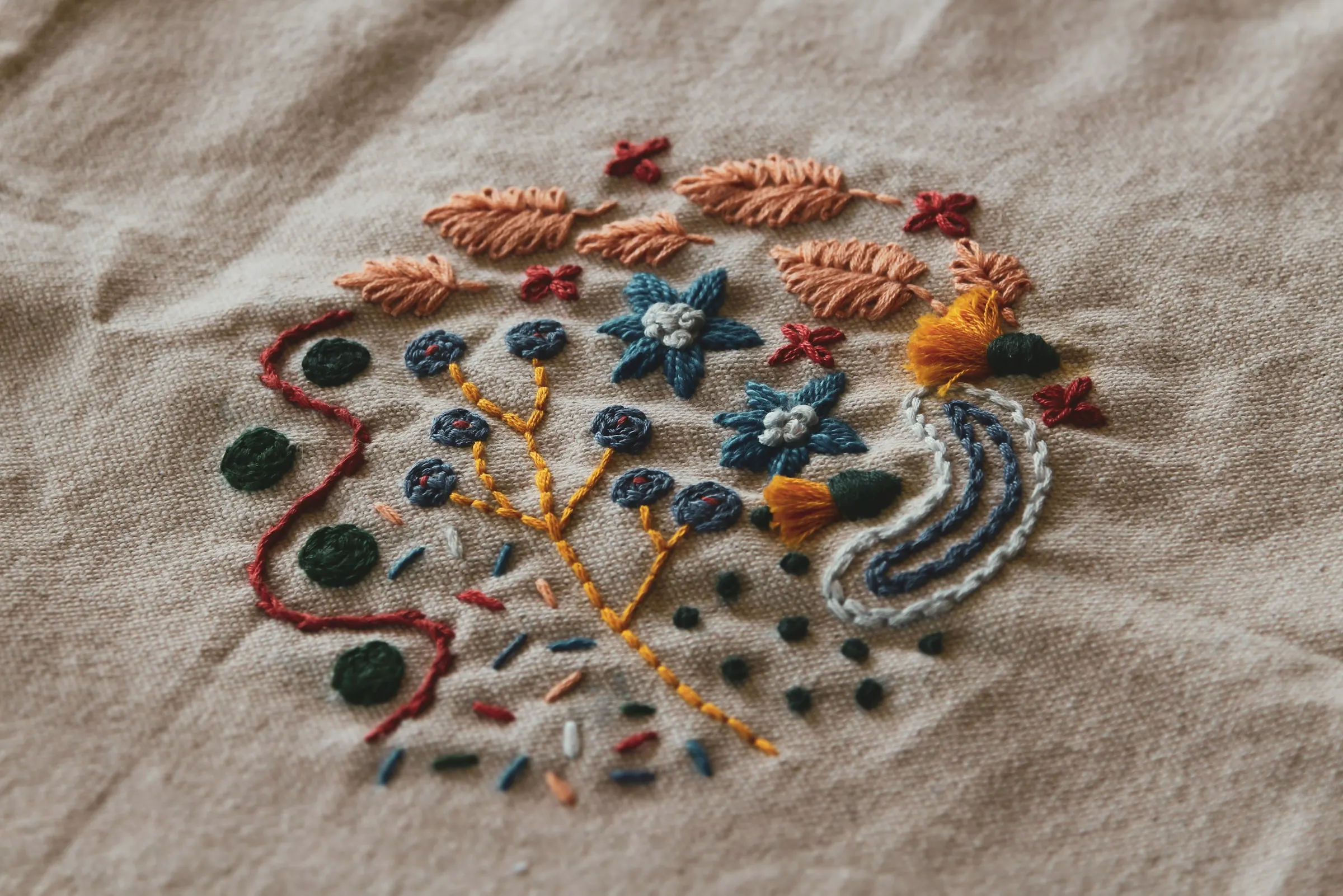 Close-up of playful hand embroidery on natural cotton: coral leaves, blue flowers, a mustard branch and scattered seed stitches