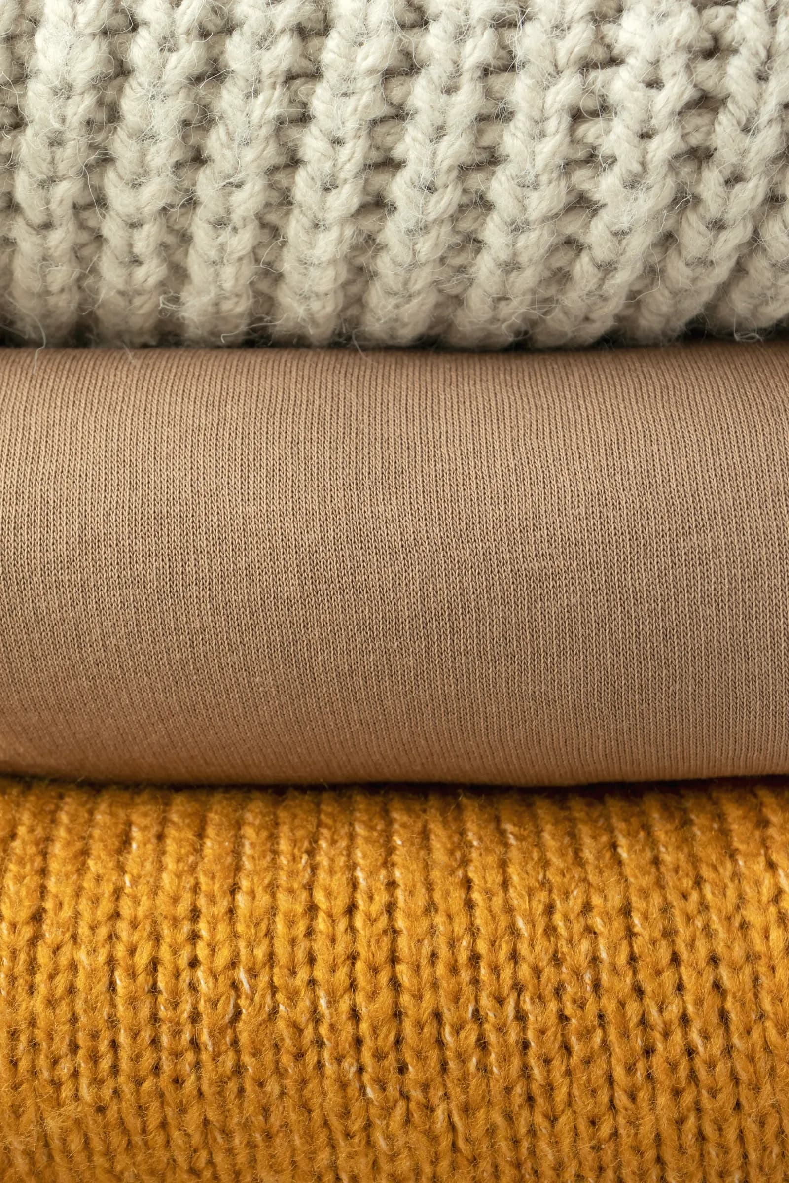 Three folded knits stacked: cream chunky rib, camel jersey and mustard sweater knit
