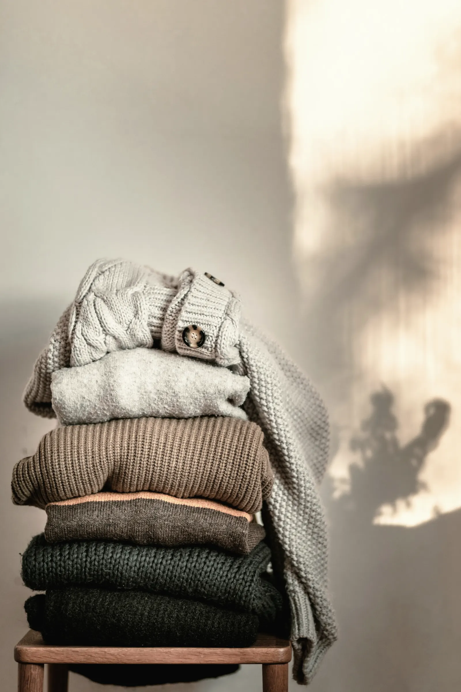 A stack of chunky knits in cream, grey, brown and forest green on a wooden stool against a sunlit wall with leaf shadows