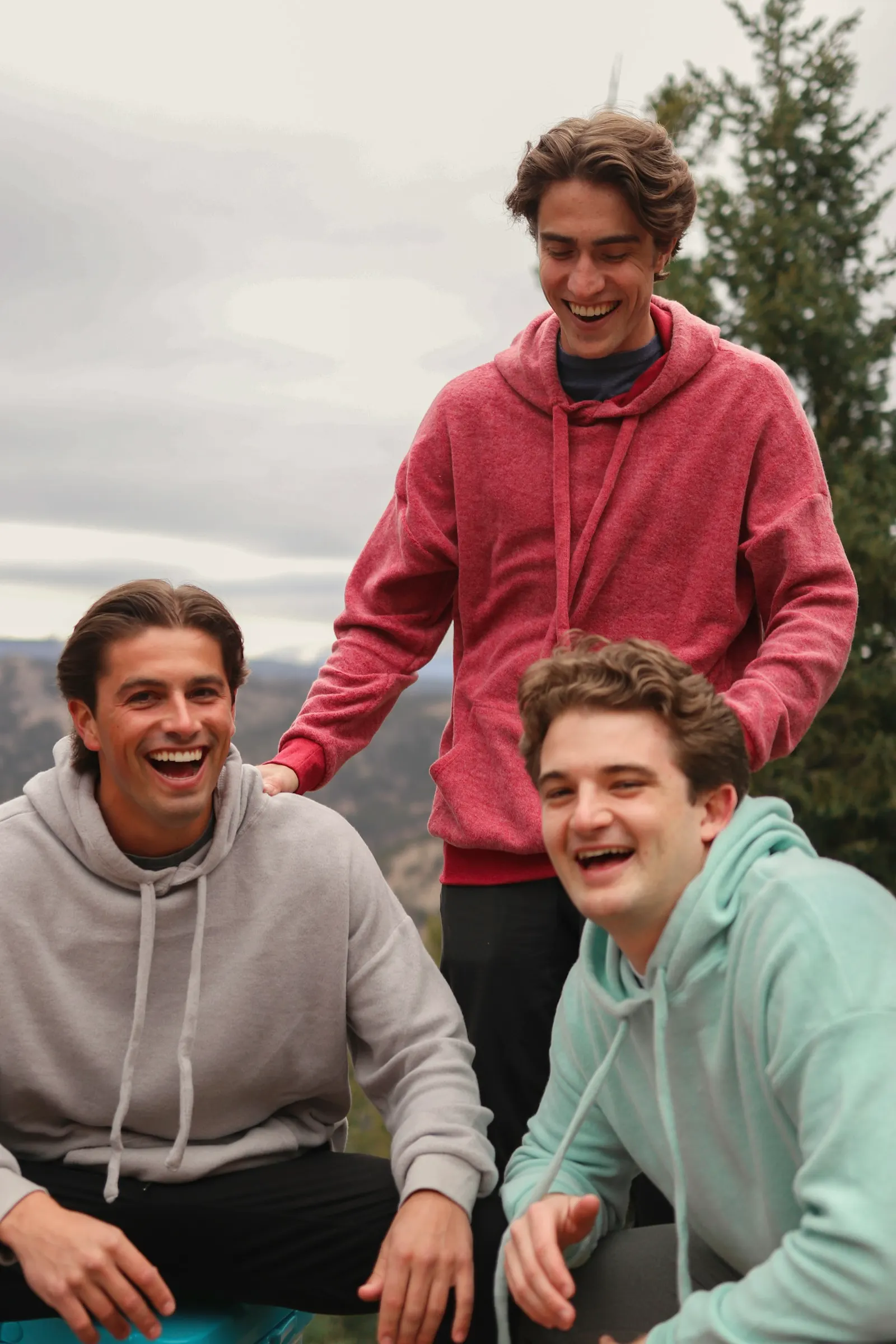Three friends laughing together on a mountain overlook in coordinated fleece hoodies in heather grey, raspberry red and mint