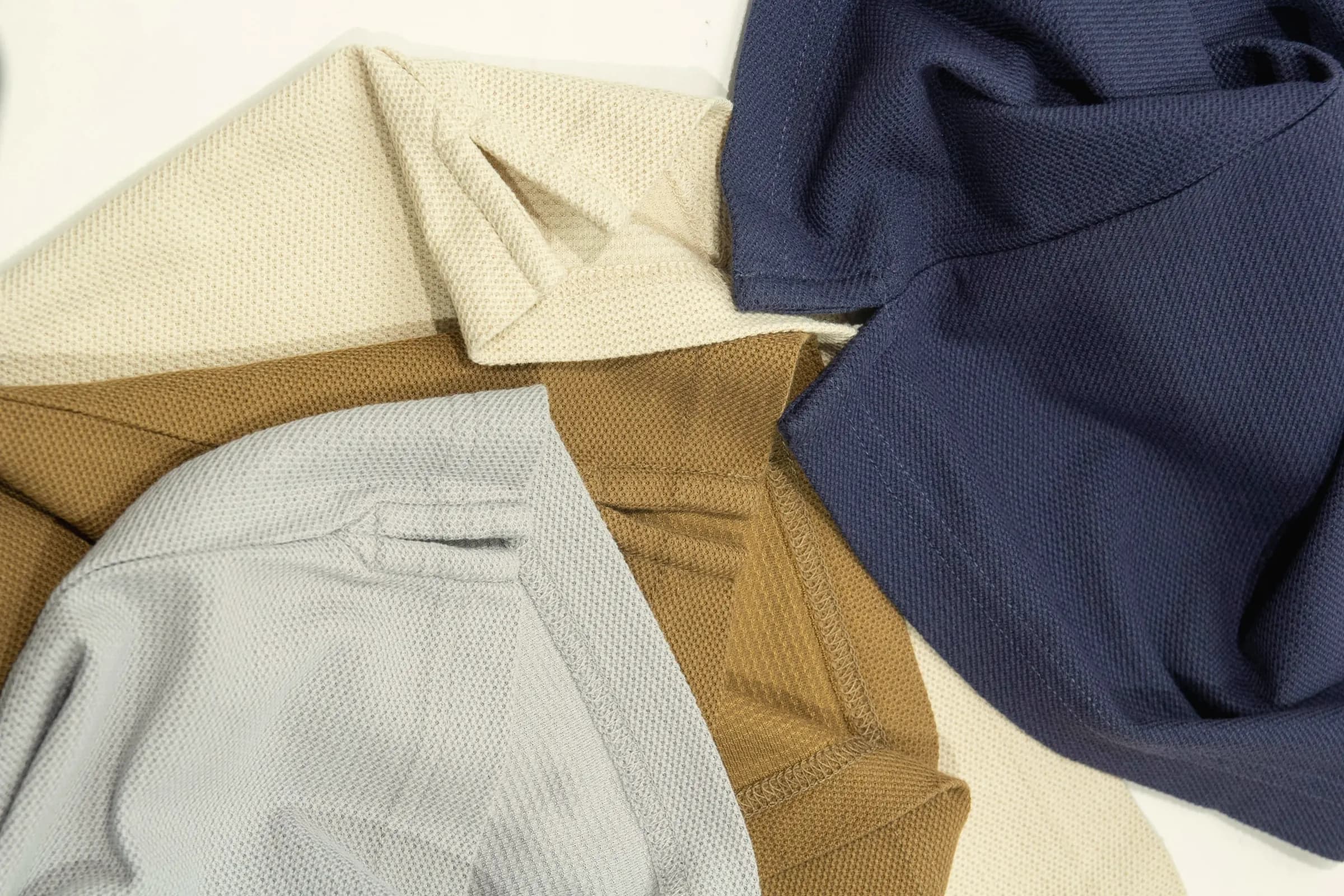 Pique knit polos in cream, camel, pale grey and navy loosely laid on a white table